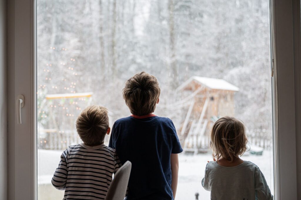 Three children gazing out a snowy scene through a glass window.