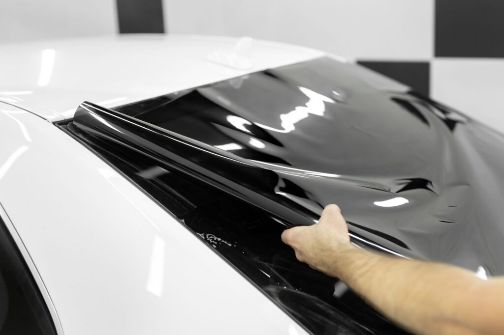 A man applies black and white window tint to a car hood, preparing for summer heat and enhancing vehicle aesthetics.