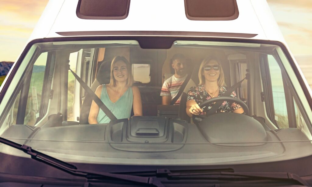 Three individuals inside a camper van, driving along the road, showcasing a broken windshield in need of replacement.